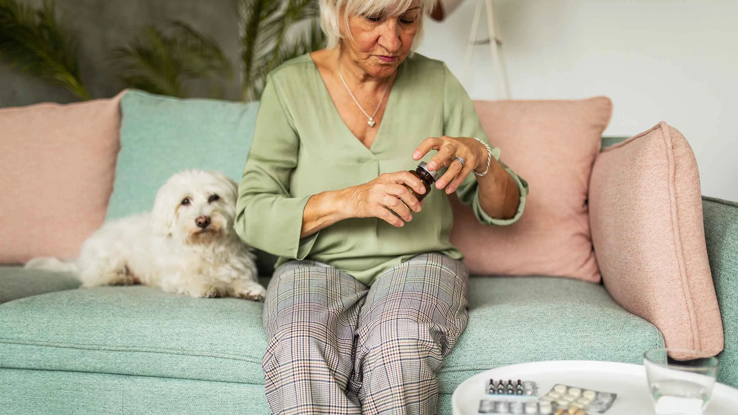 Senior woman opening her medicine bottle while sitting on the couch next to her dog.