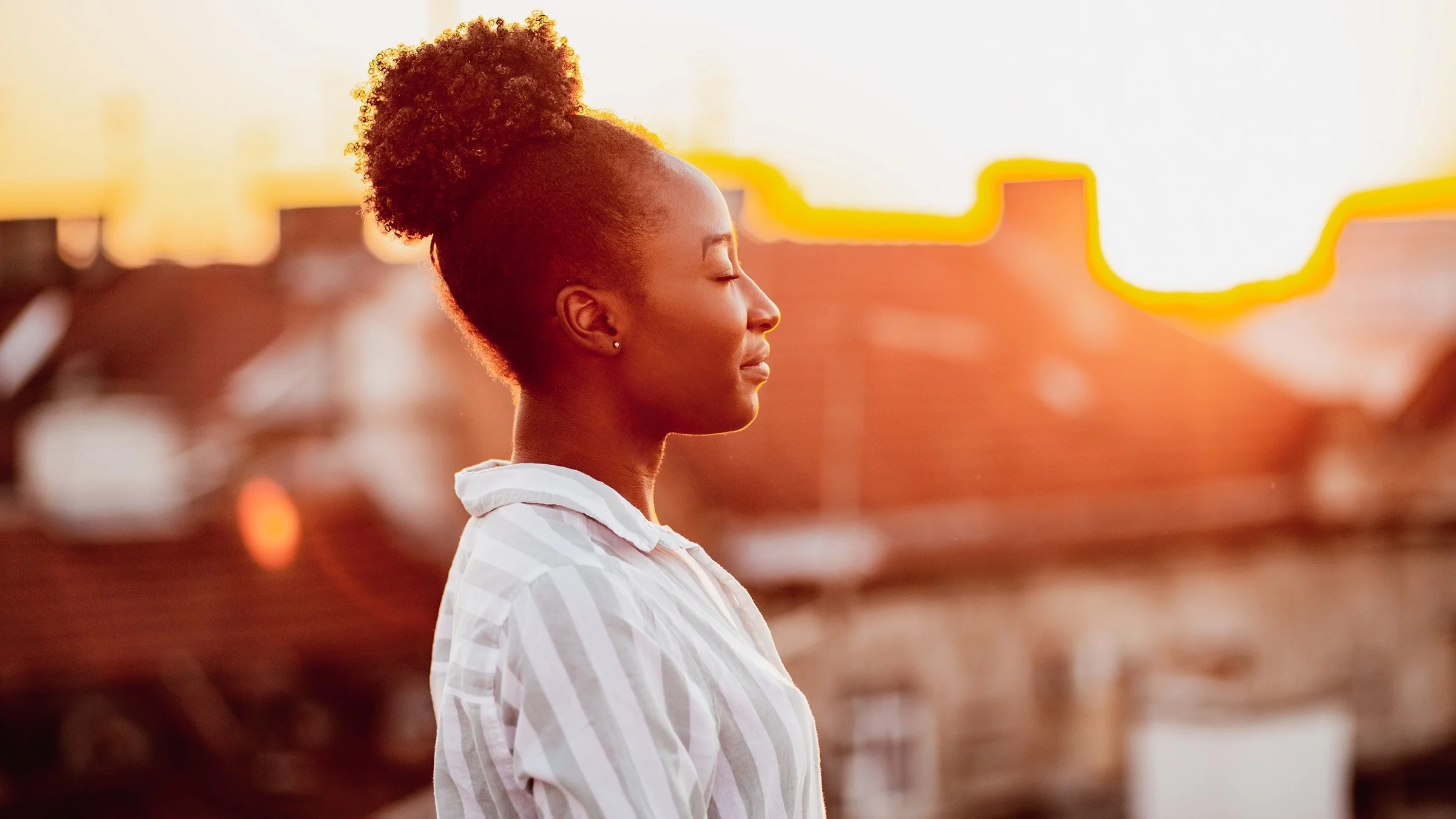 Young Black woman closing her eyes and meditating outside during sunset on a rooftop terrace.