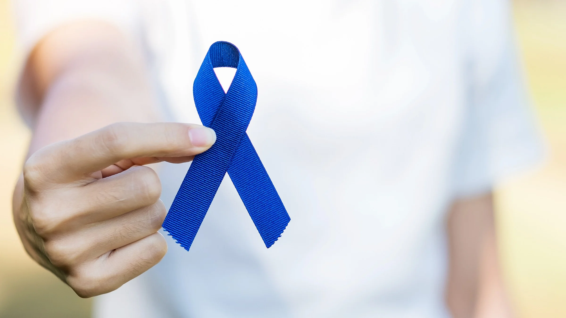Close-up of a person holding a dark blue cause ribbon for Colorectal Cancer Awareness month. They are standing outside with greenery as the background.