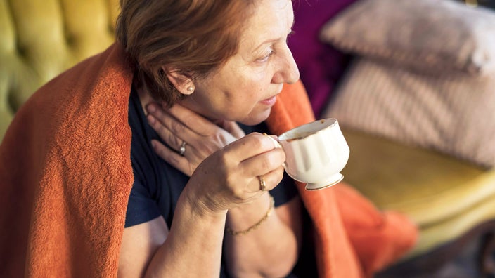 A woman with a sore throat is on the couch in a blanket.