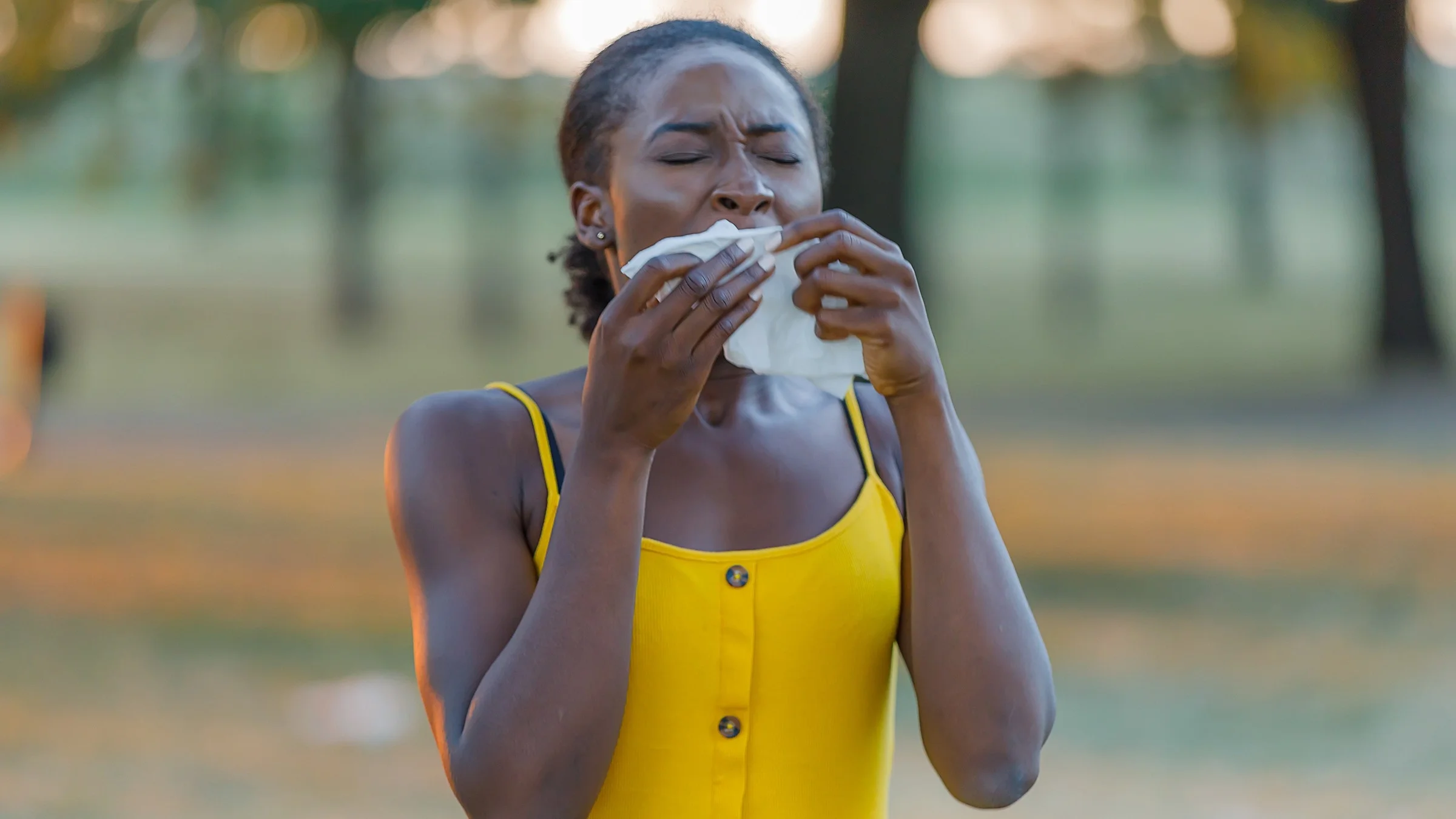 Portrait of a woman outside sneezing into a tissue. She is wearing a yellow tank top.