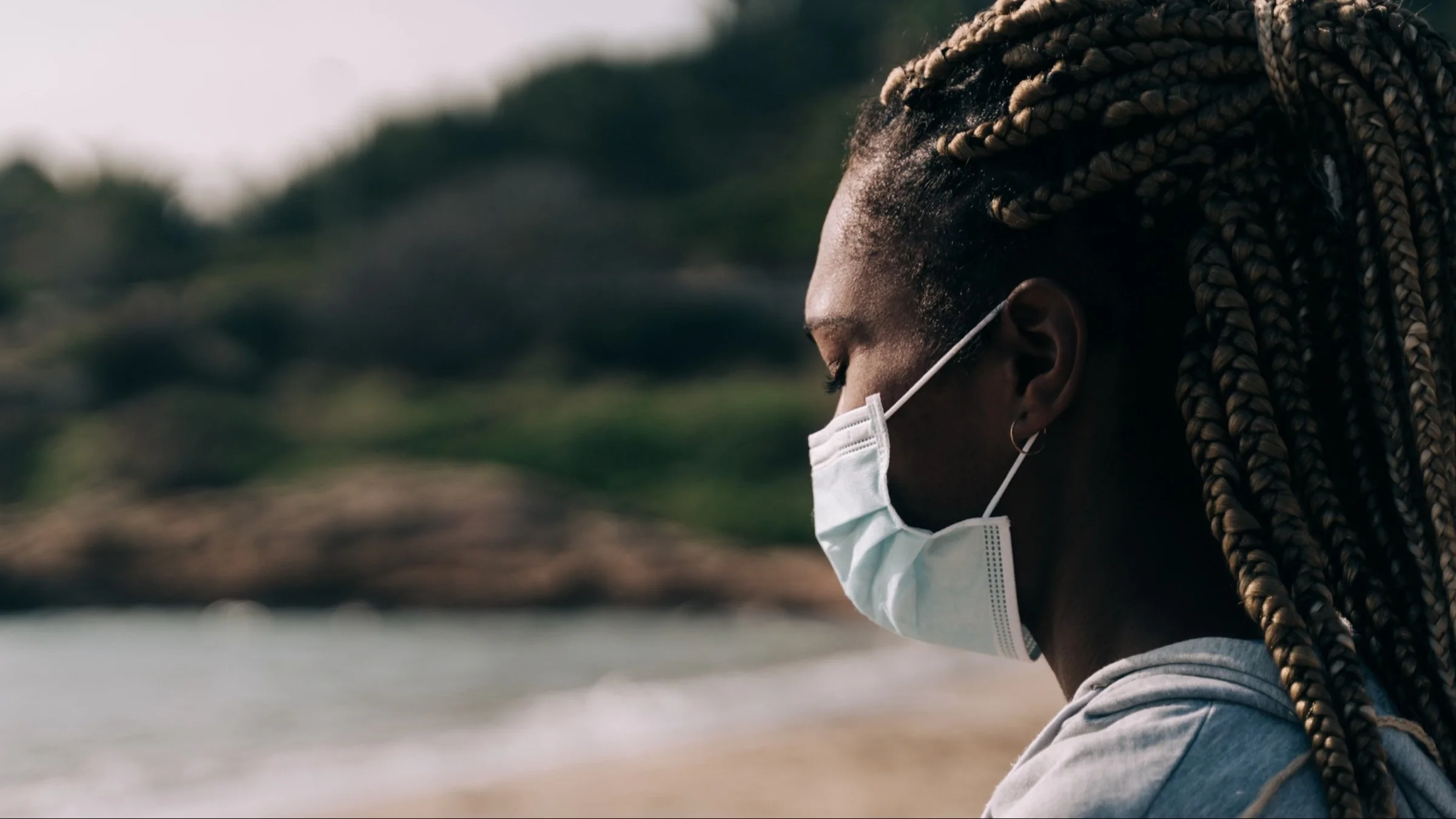 Side portrait of a woman at the beach wearing a medical face mask looking down and sad.
