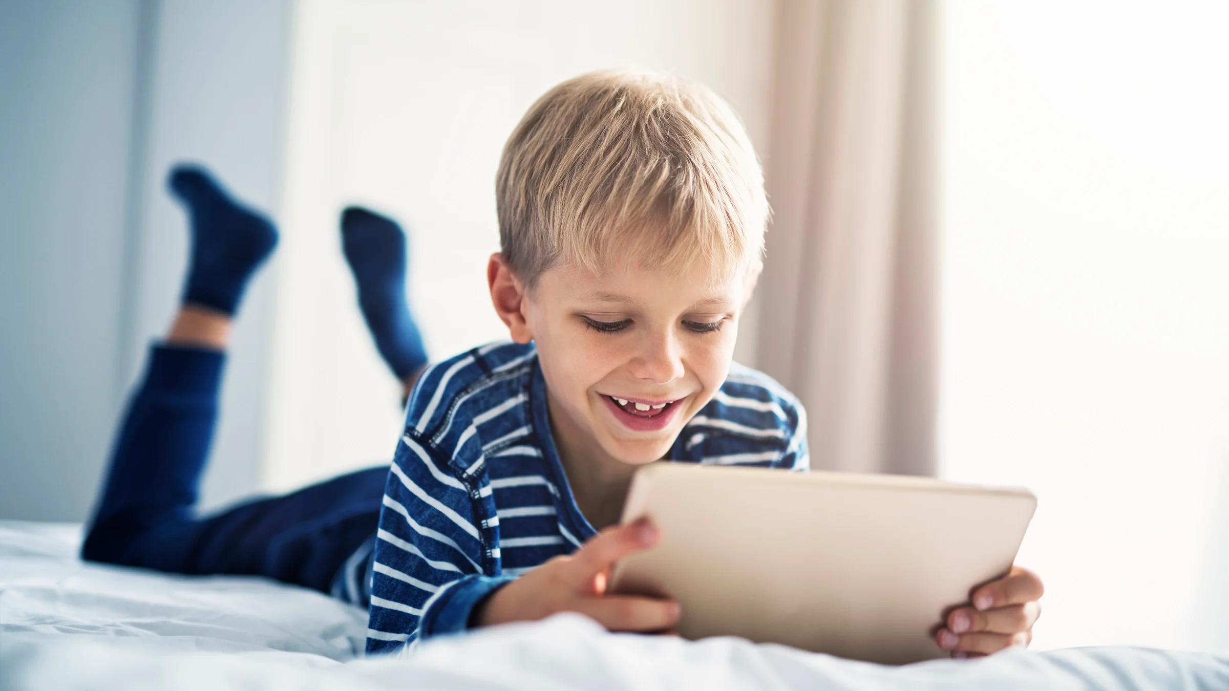 Blonde child smiling while he is playing games on his tablet.