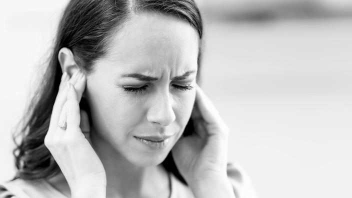 Black and white close-up of a woman with ringing in her ears. She is covering her ears with her hands and is squinting in pain.