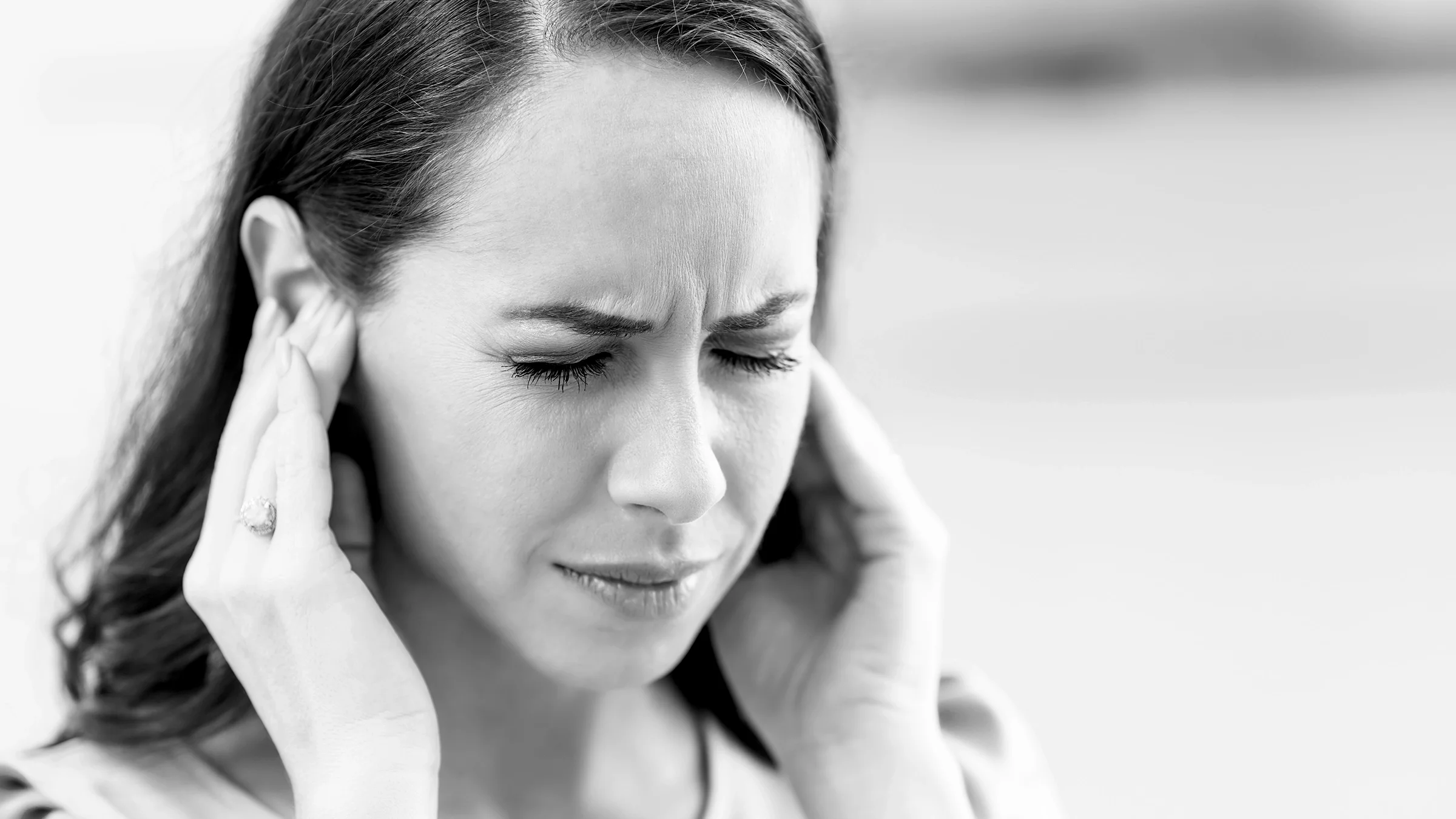 Black and white close-up of a woman with ringing in her ears. She is covering her ears with her hands and is squinting in pain.