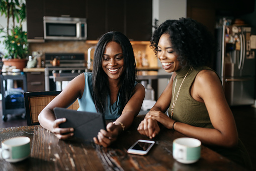 Two friends looking at a tablet and smiling while having coffe at the kitchen table.