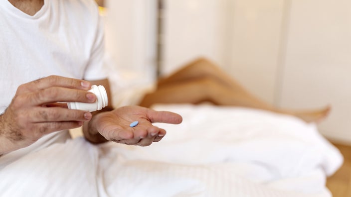 A man prepares to take a pill that’s resting in the palm of his hand. In the background, a woman lies on a bed out of focus.
