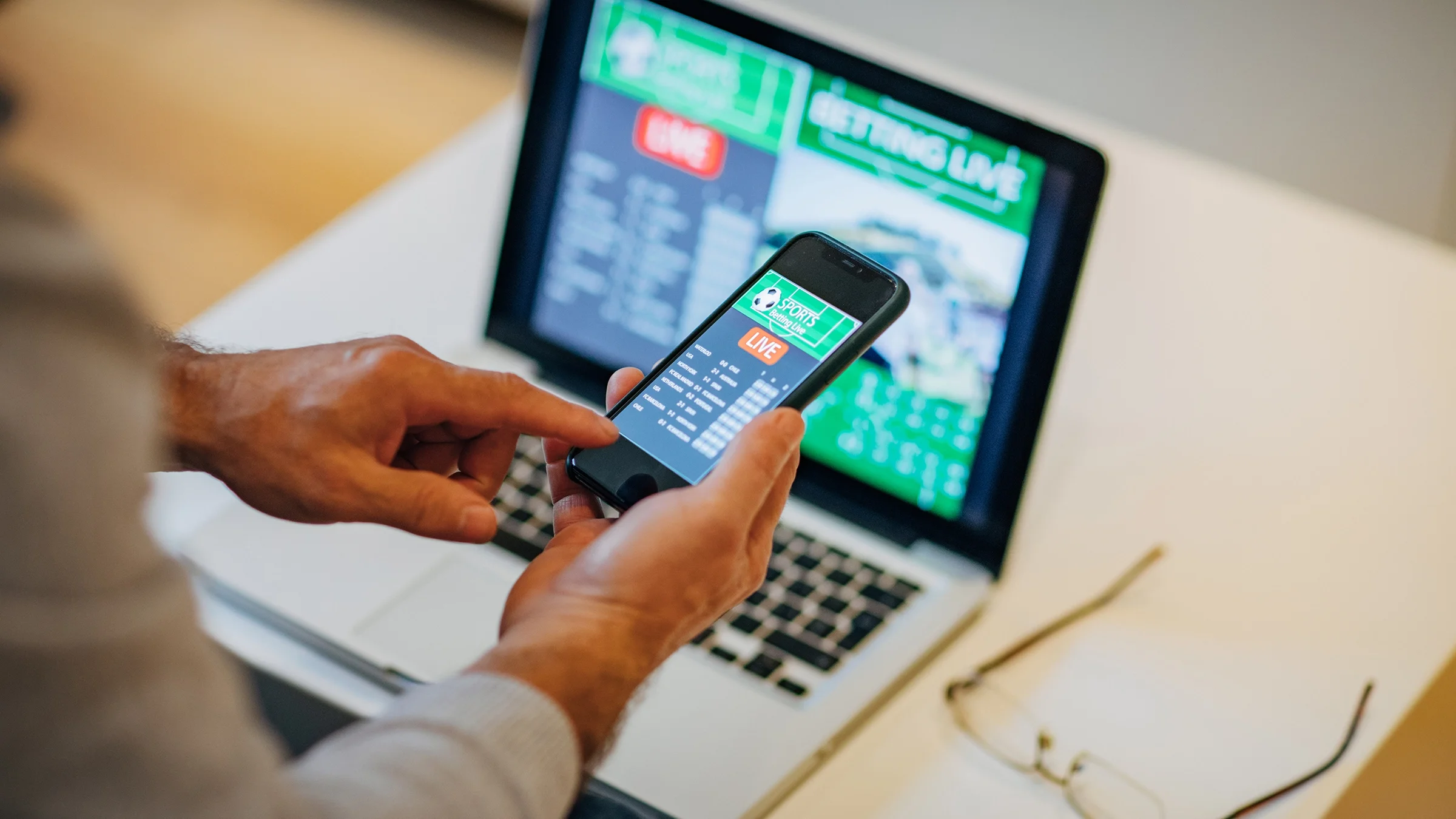 Close-up person holding mobile phone with sports gambling page up. In the background there is a laptop with gambling pages up too.