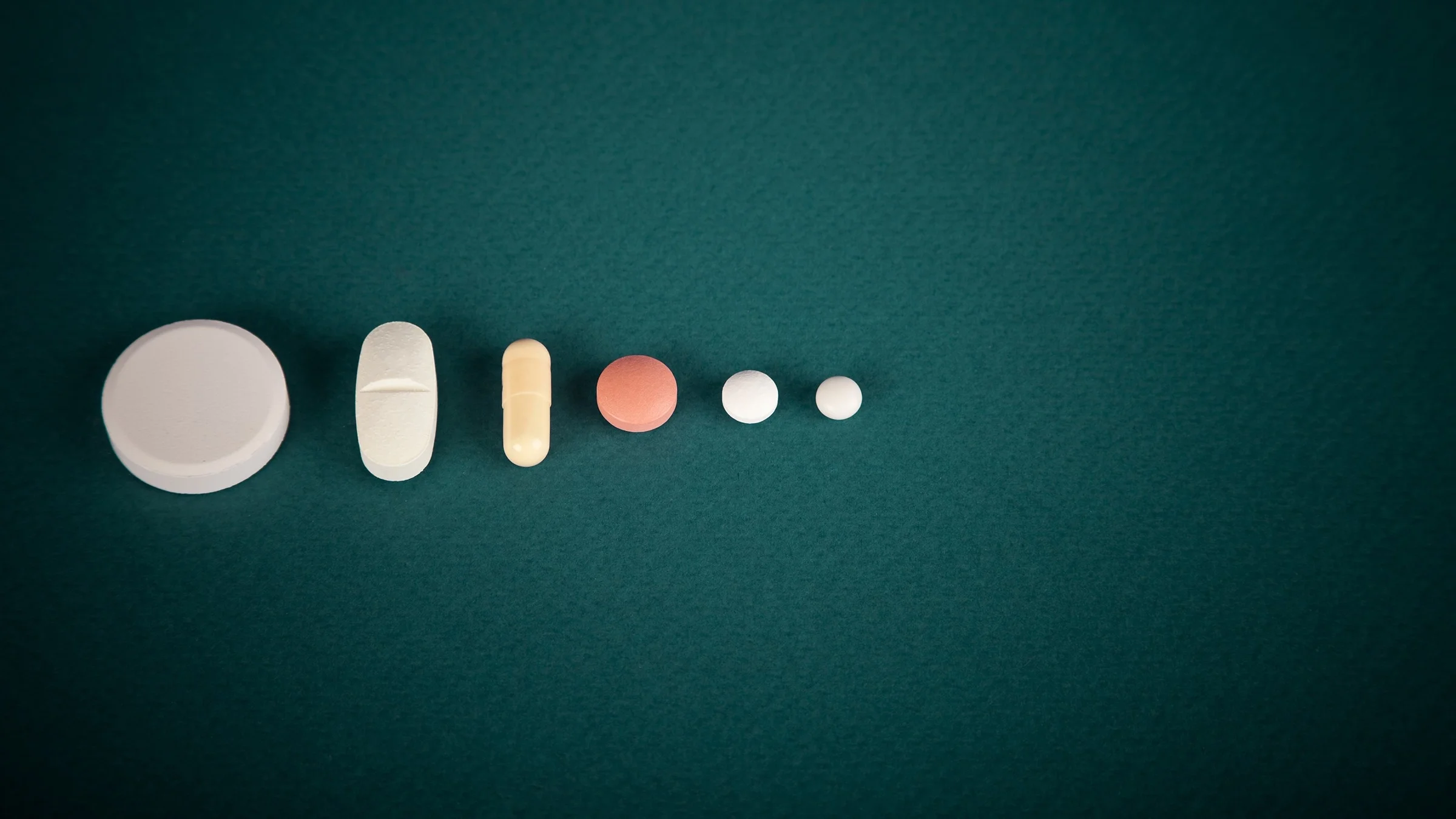 Various pills lined up from largest to smallest in a line from left to right on a dark green-blue background.