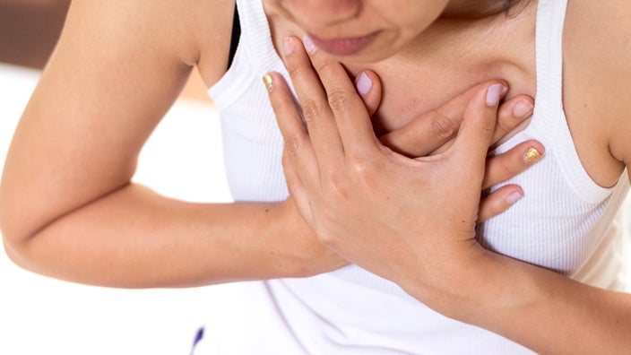 Cropped shot of a woman clutching her chest in pain. She is wearing a white tank top and has pink and gold nail polish.