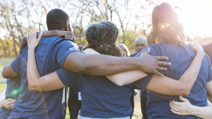 A group of people circling up with their arms over each other’s shoulders. They are outdoors at a park and the focus is on their backs.