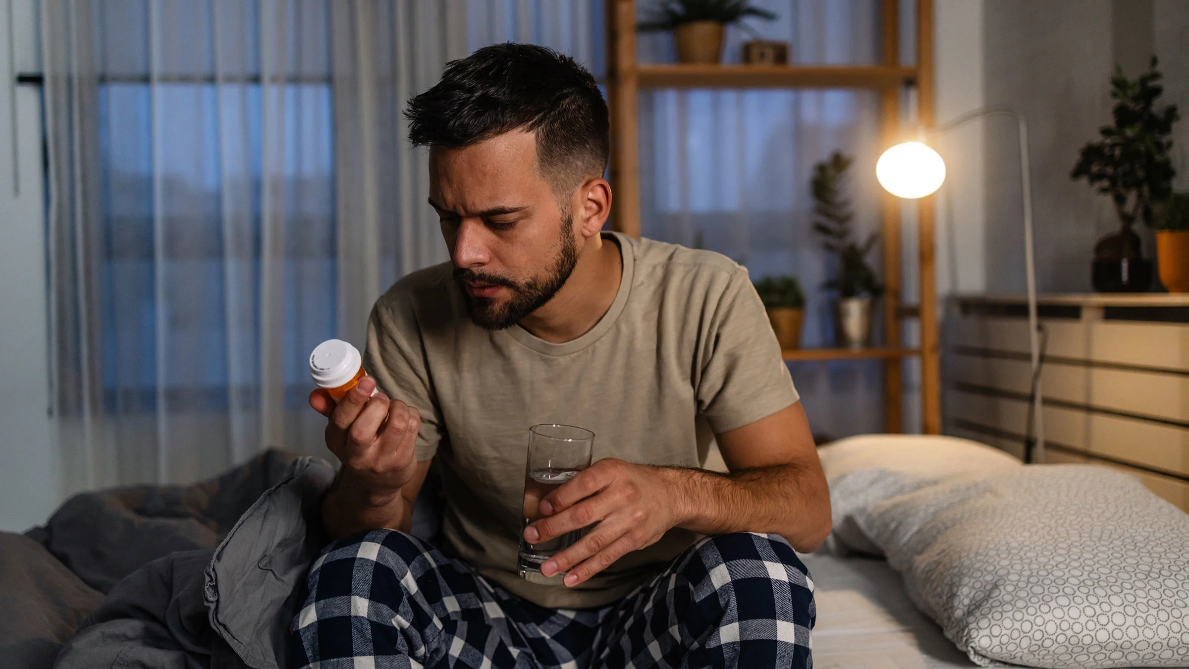 A man reads a pill bottle label while sitting on his bed.