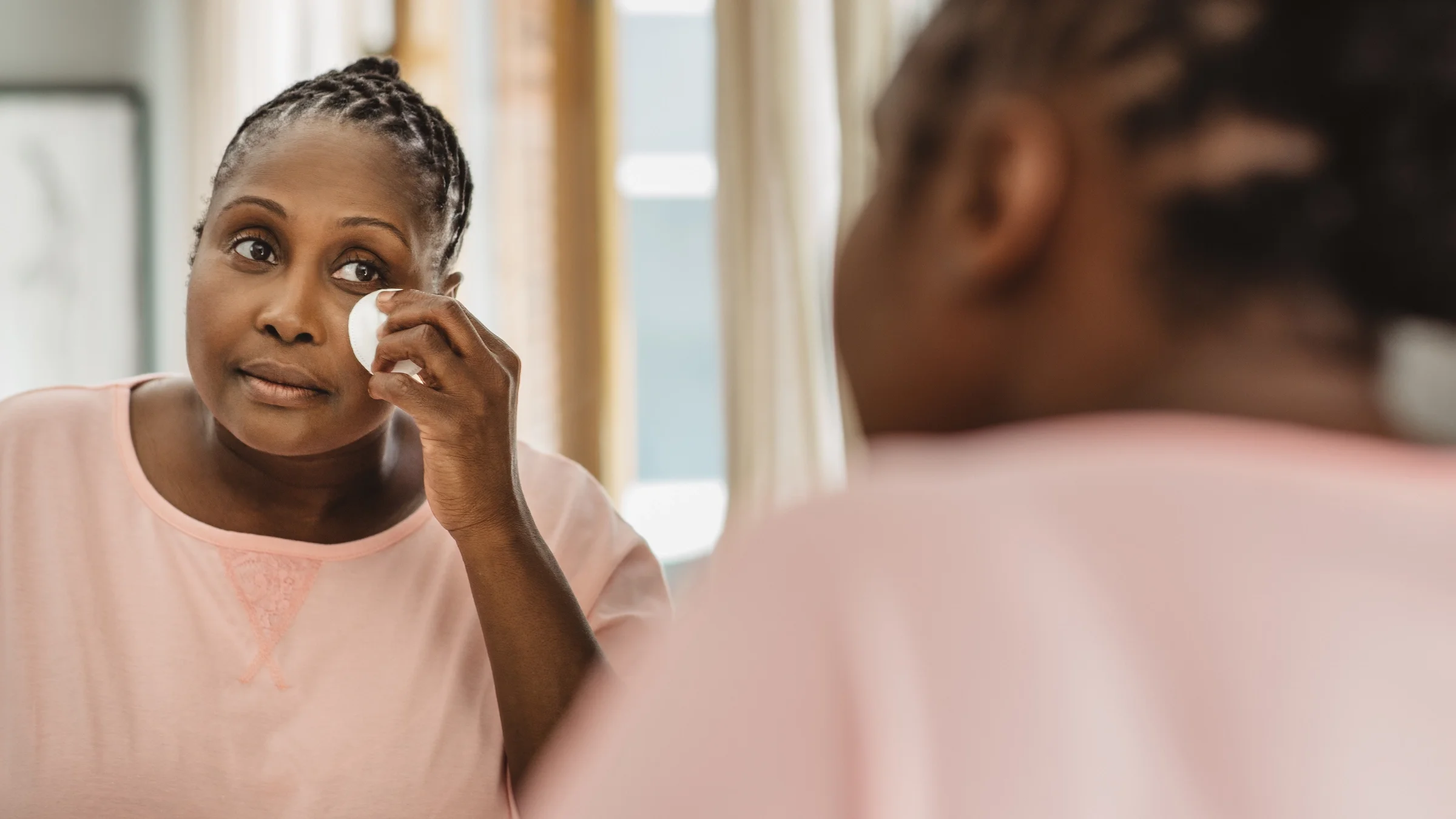 A woman uses a cotton pad to clean her face while looking in a bathroom mirror