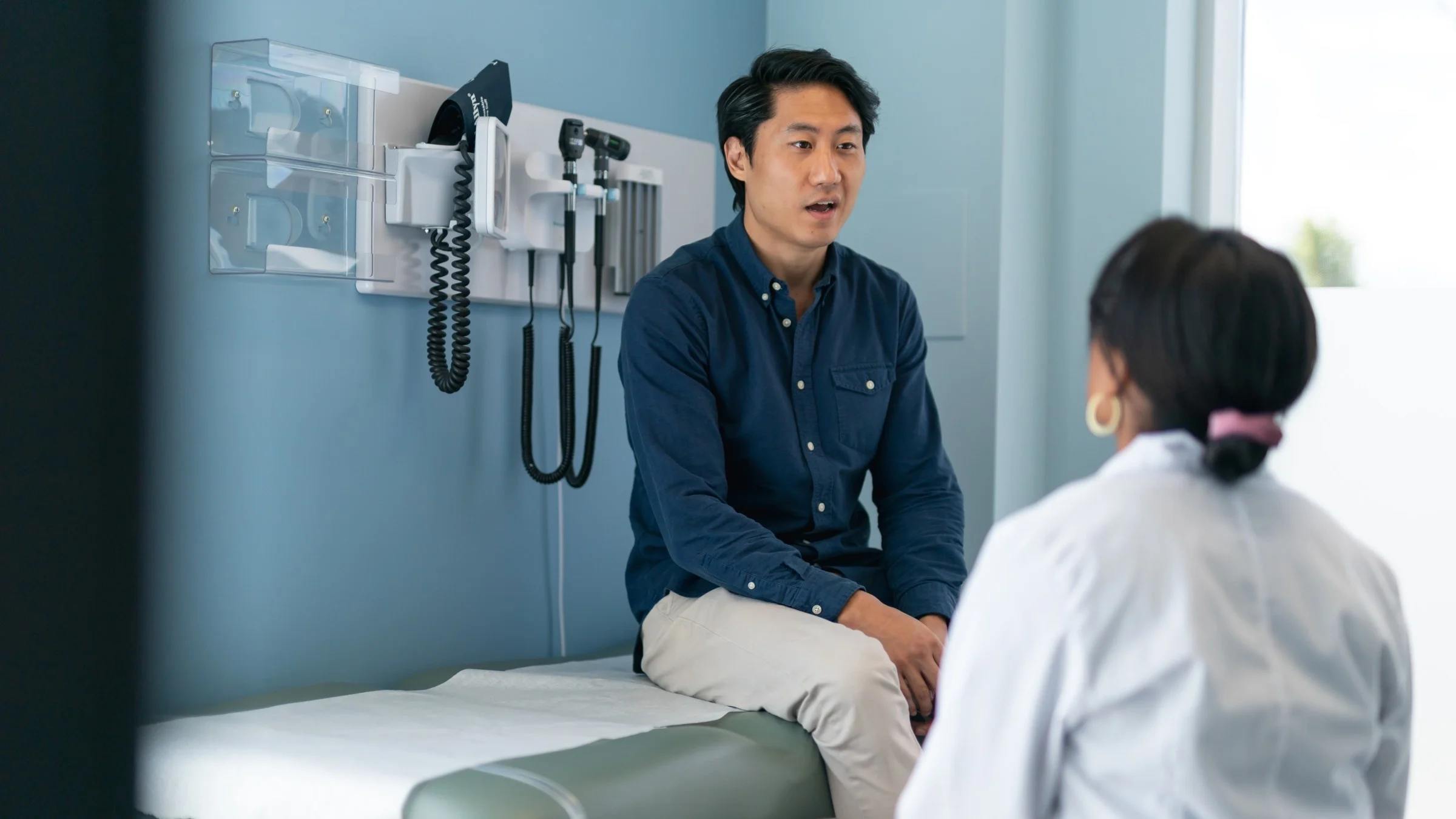 A man sitting on the exam table in the doctors office talking to his doctor sitting lower than him.