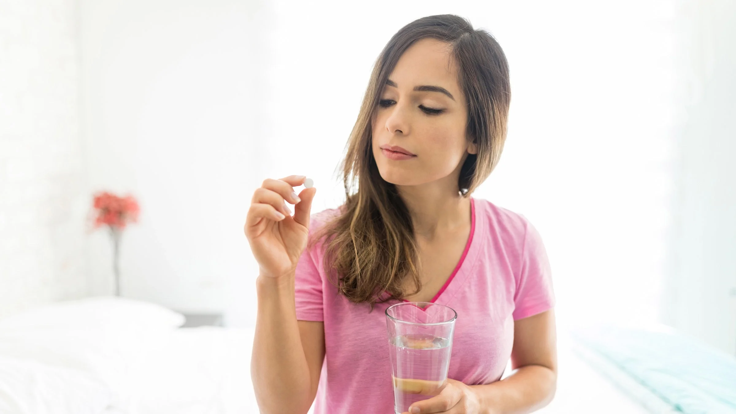A woman sitting on a bed holding a glass of water and looking at a medicine tablet.