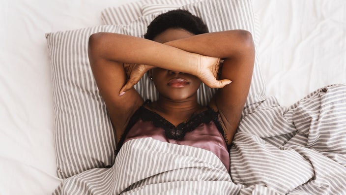 Young woman who can't sleep with her arms crossed over her eyes laying in bed with striped sheets.