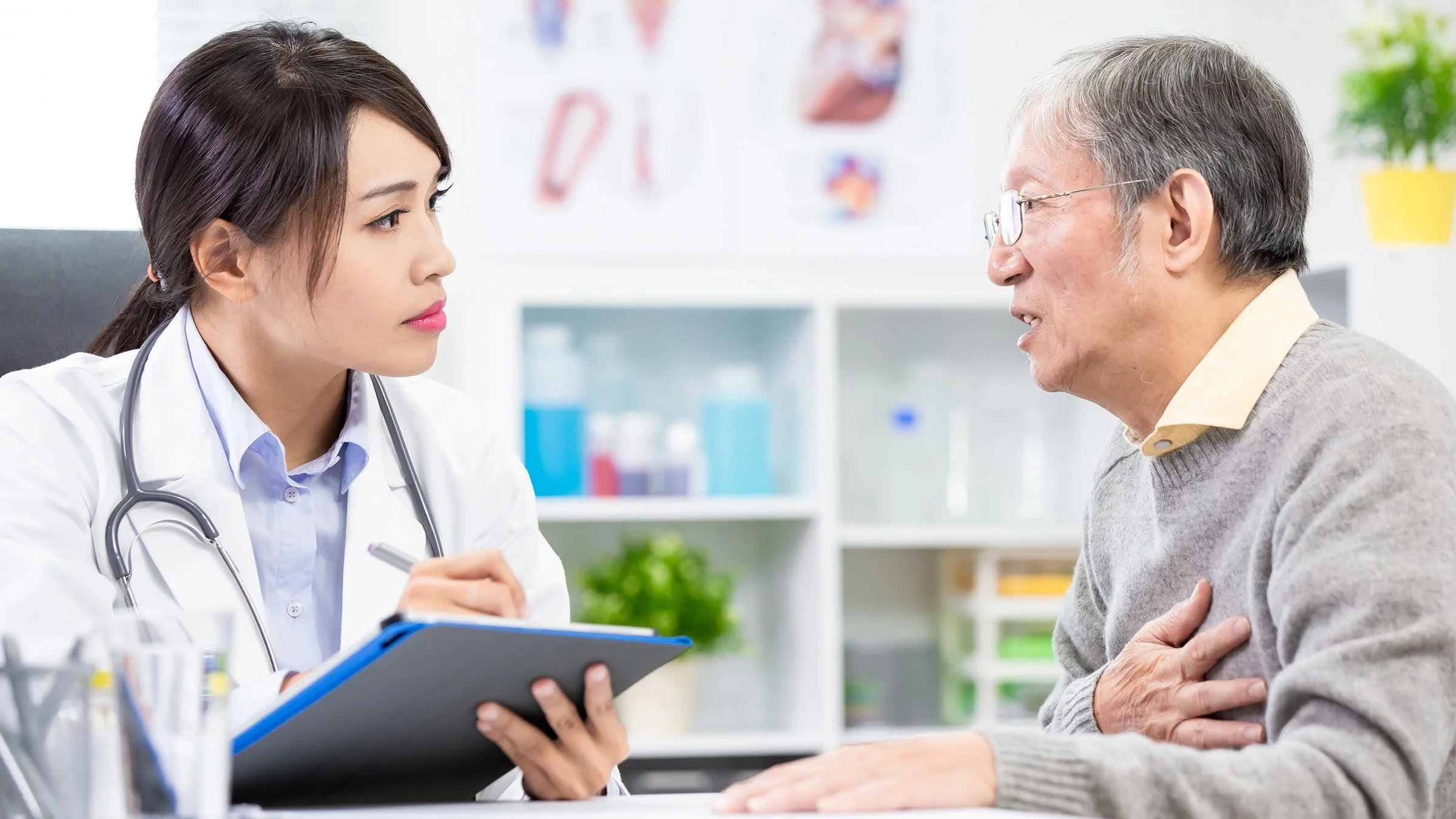 A older patient clutching their chest, while talking to a doctor with a clipboard.