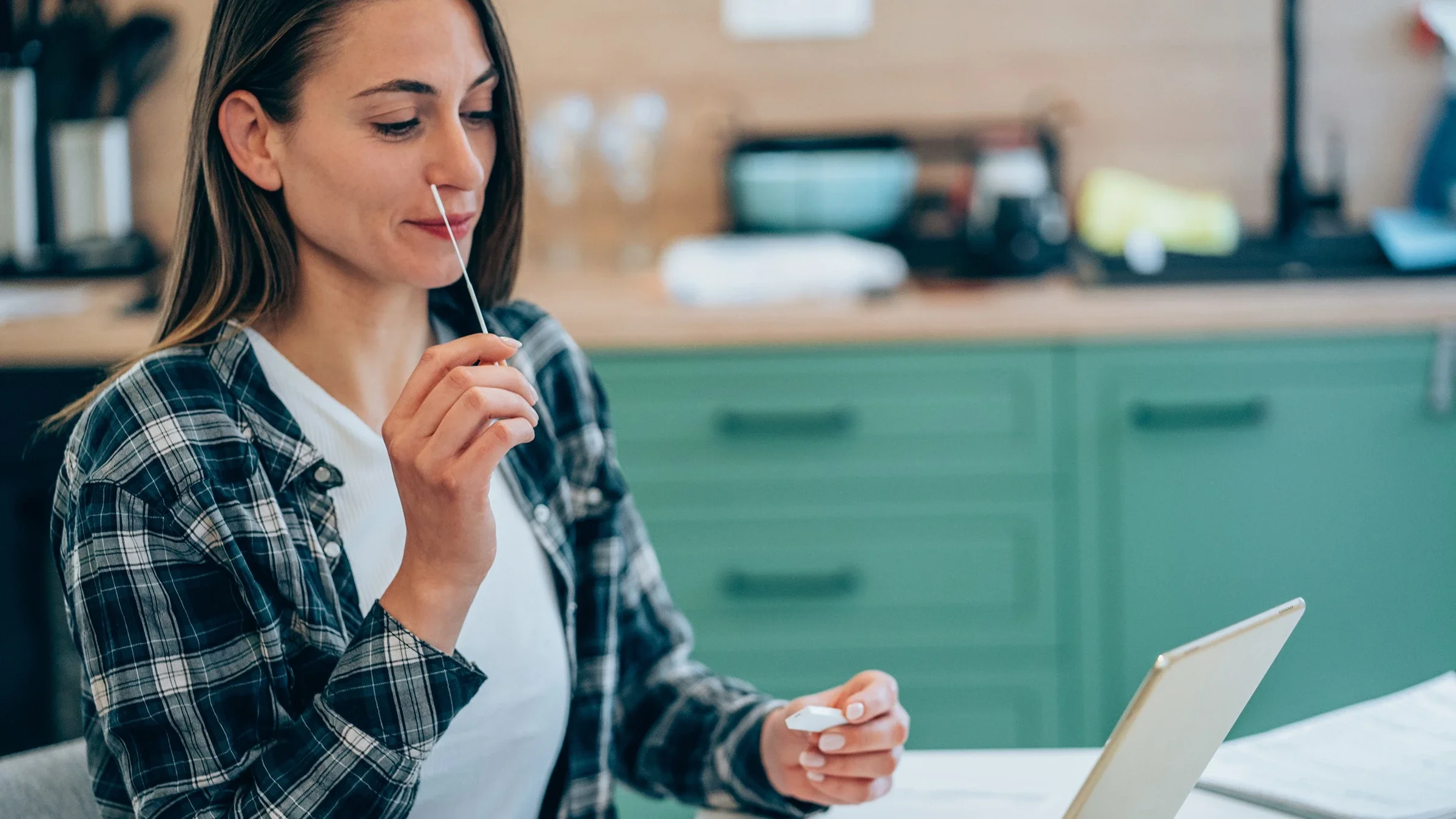 Young woman using an at home COVID test. She is sitting at the dining table with her tablet propped up in front of her.