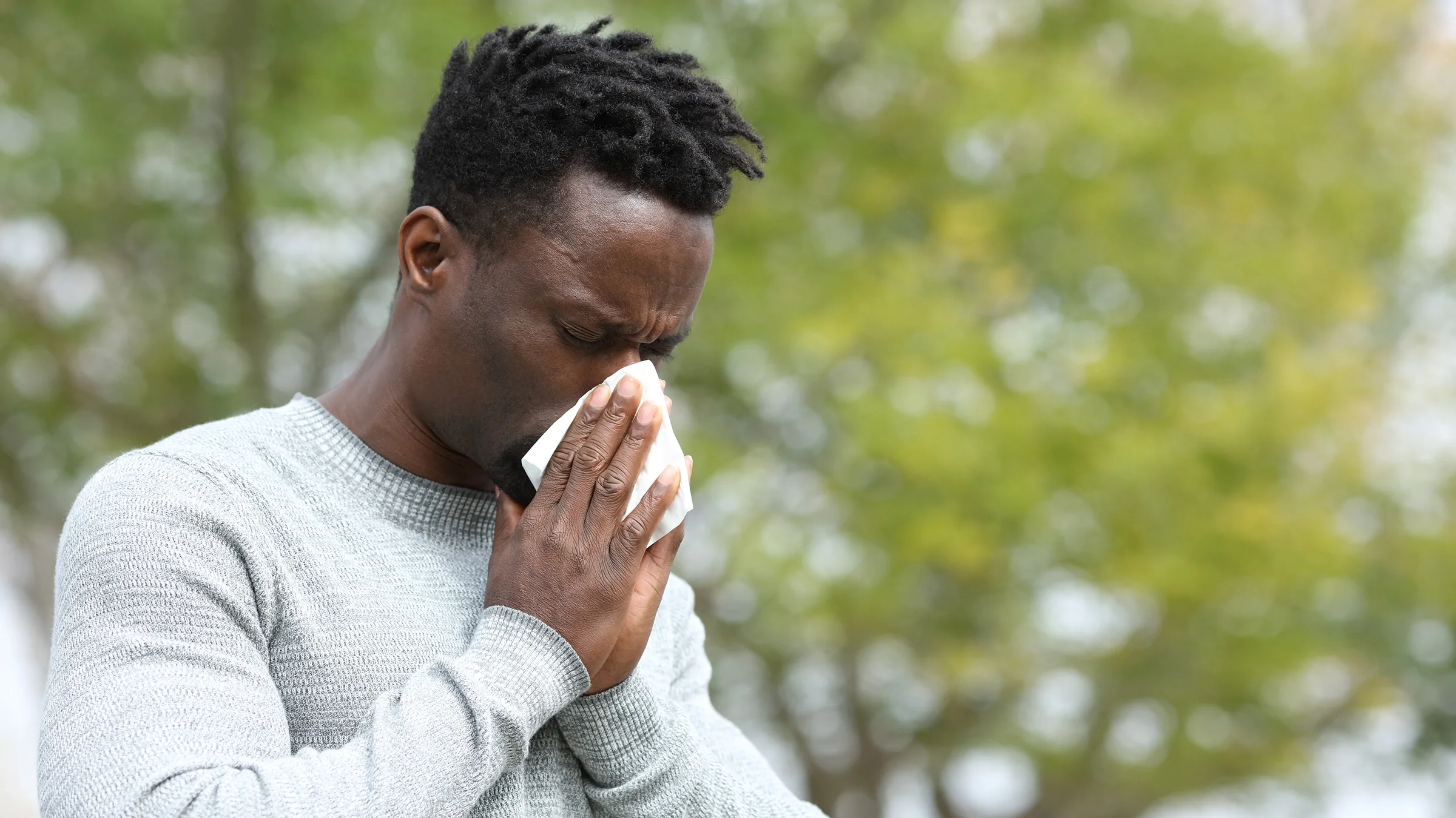 Young man sneezing into a tissue while he is outside in the park.
