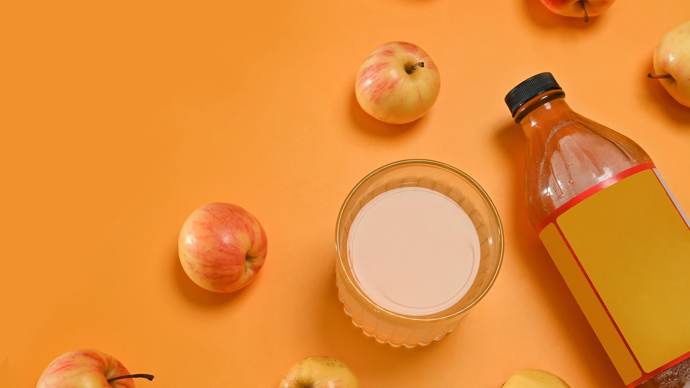 Glass of apple cider vinegar styled with a bottle and apples on an orange background.