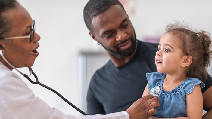 Father holding his young daughter while the doctor examines her with a stethoscope.