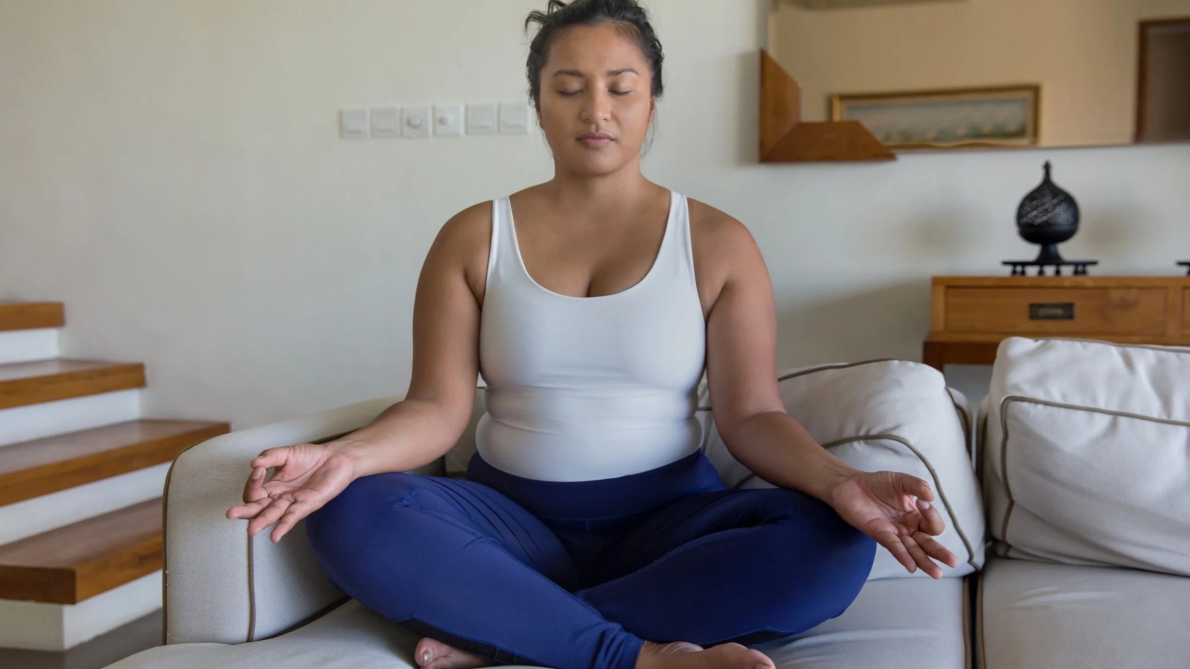 Young woman sitting cross legged on the couch with her eyes closed meditating and doing breathing exercises.