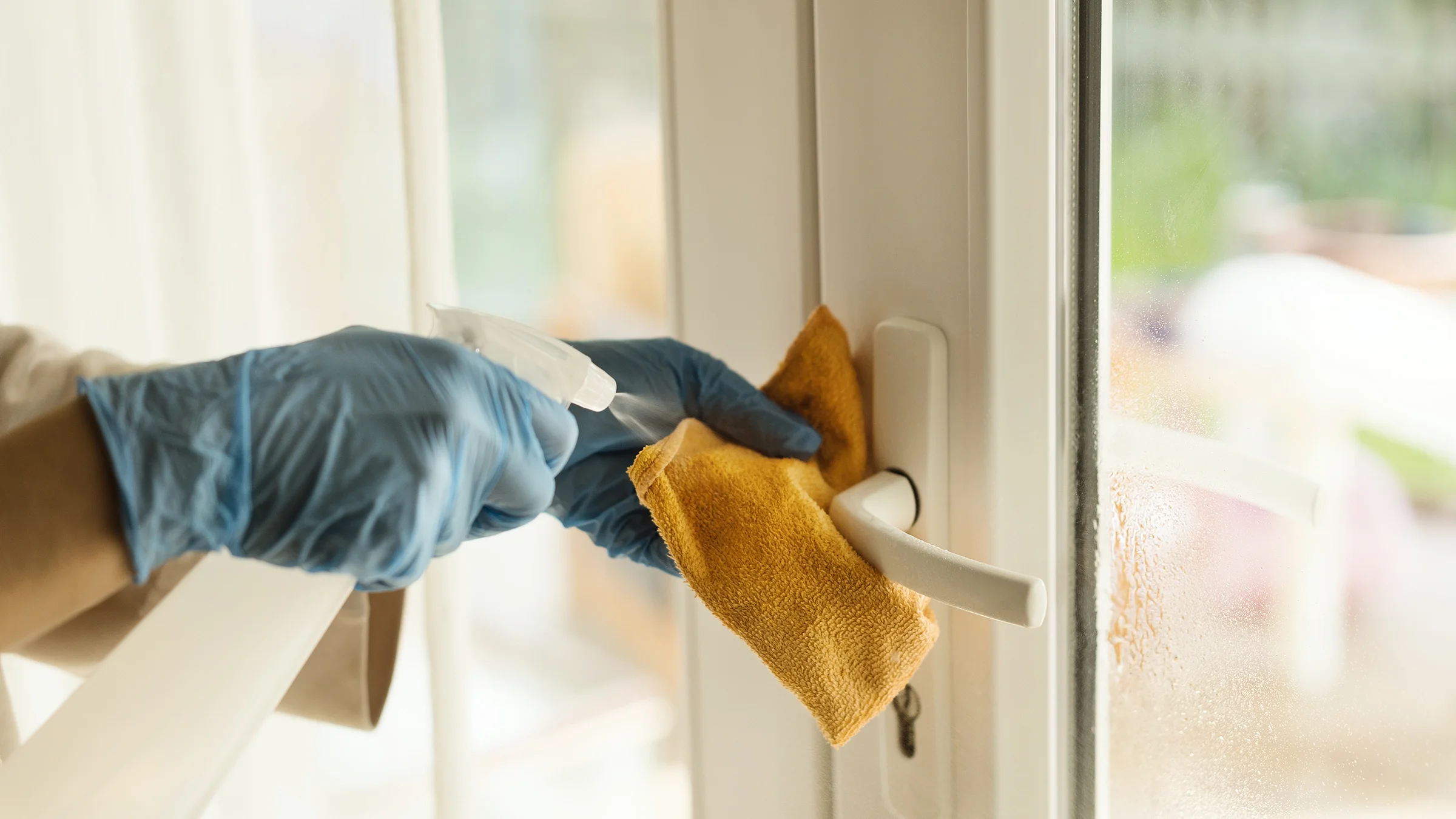 Close up of a person cleaning a door white door handle with blue gloves, orange cloth, and cleaning spray.