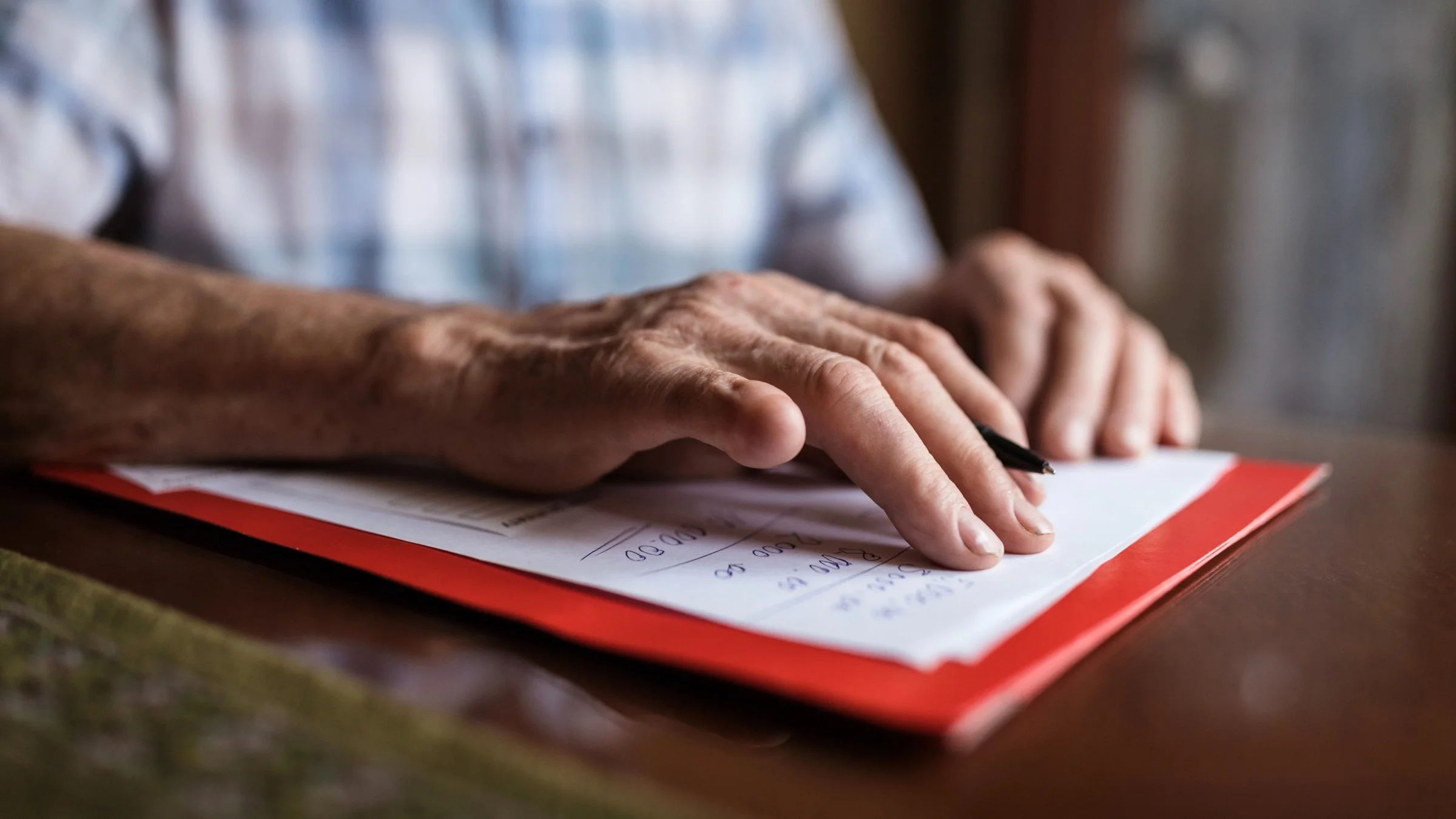 Close-up of an elderly mans hands reviewing his written medical expenses on paper on top of a red folder.