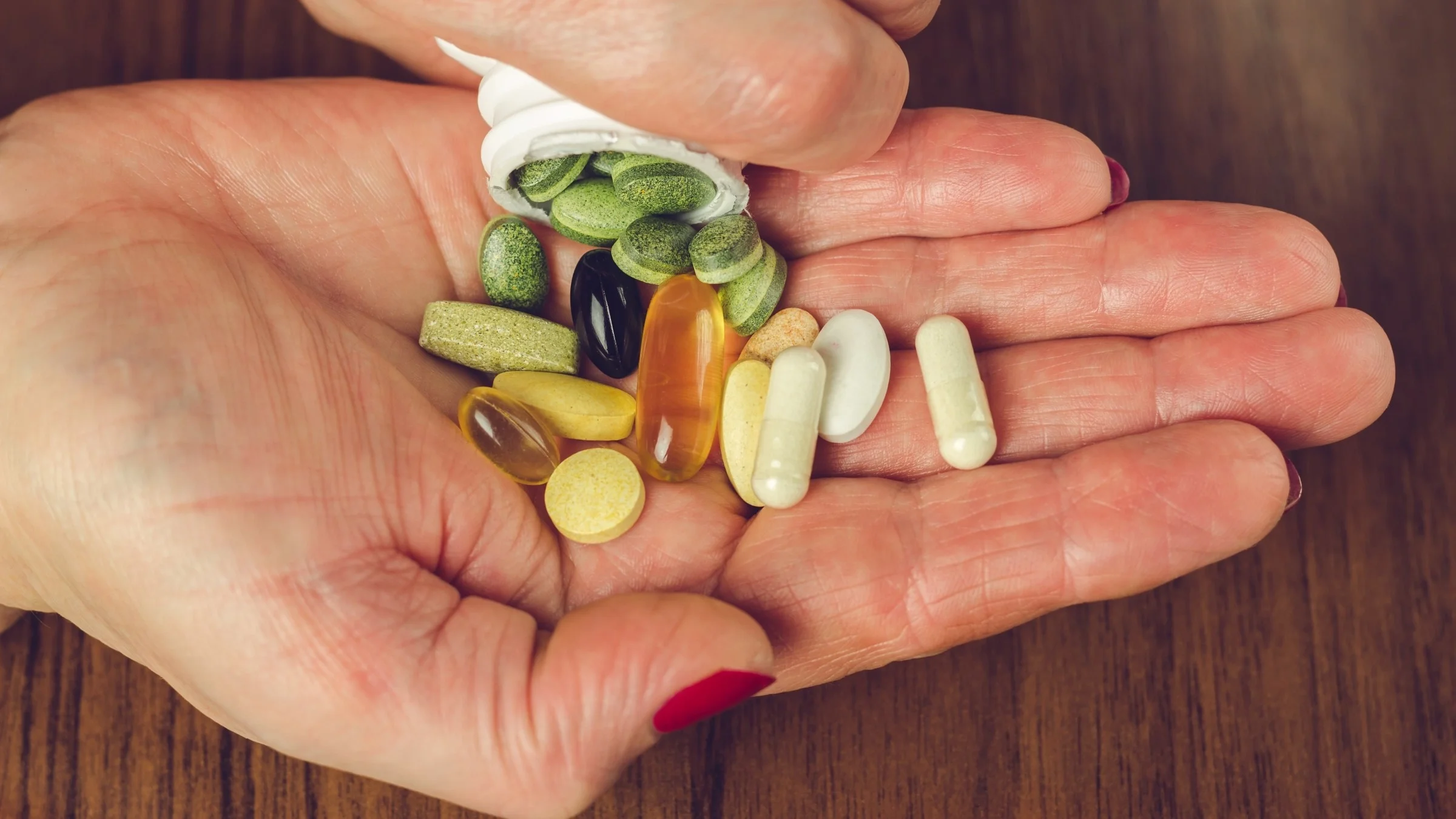 Close-up on a person's hands pouring a variety of supplements of different shapes, sizes, and colors onto their hand on a textured wood background.