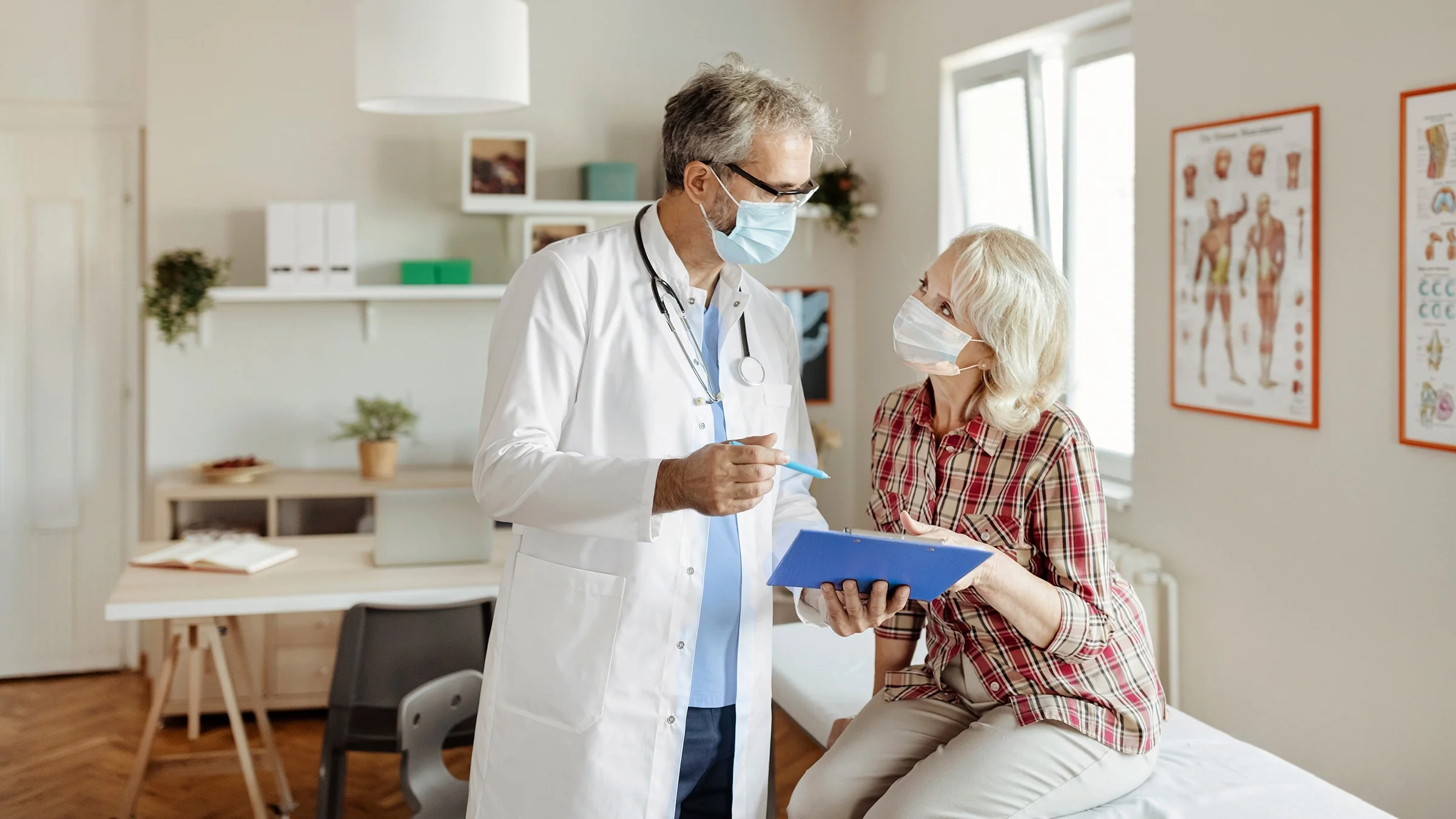 Older woman patient going over her chart with her doctor in the exam room. Both are wearing blue medical face masks.