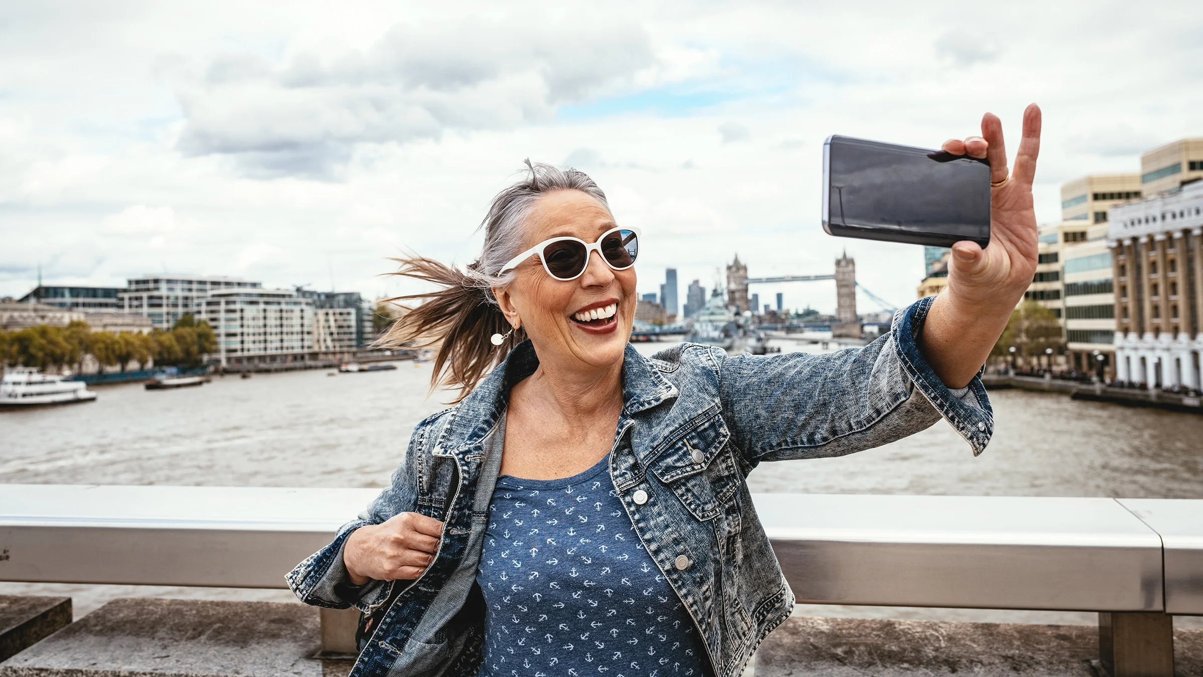 Senior woman smiling while taking a selfie next to the Thames River in London while traveling.