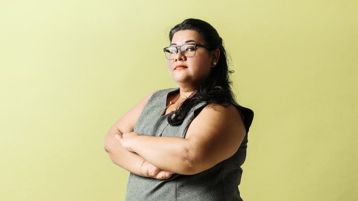 Studio portrait of a Latina woman. She is standing in a power stance and looking directly at the camera. The background is a light-yellow color.