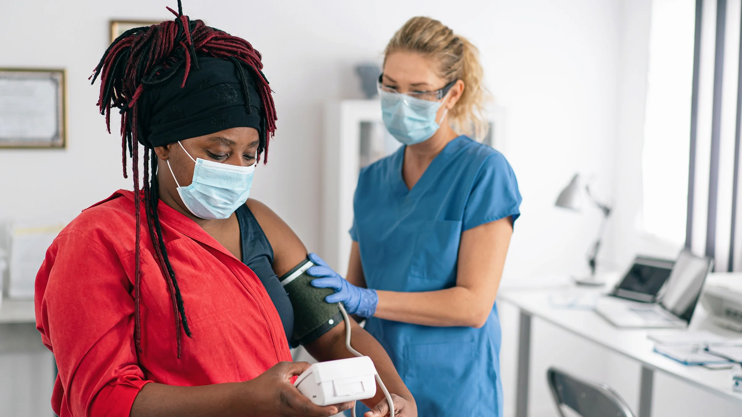Woman with braids working with her nurse to take her blood pressure using a blood pressure cuff. They are in an exam room.