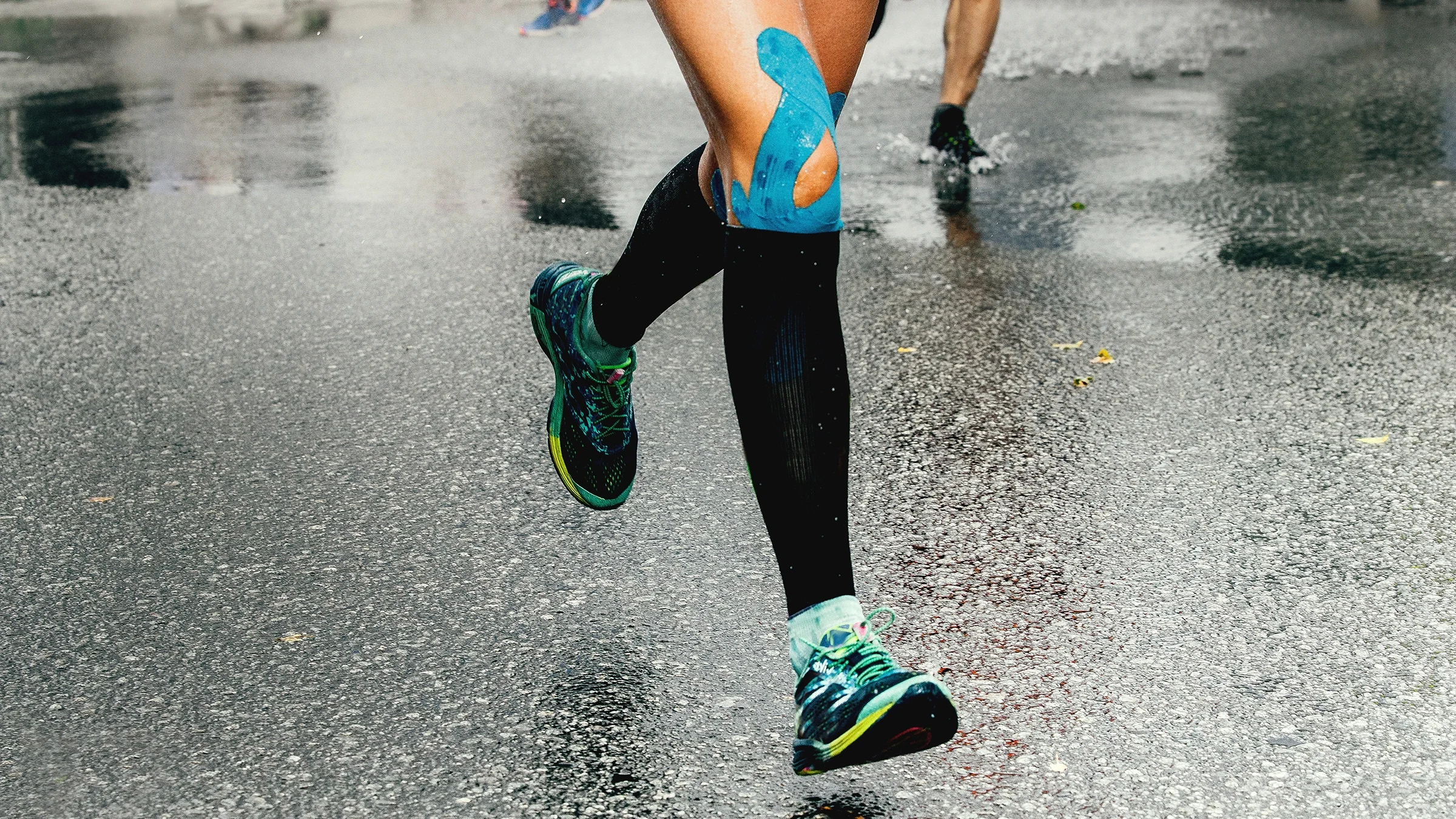 Cropped shot of a woman running on wet pavement. They are wearing high socks but you can see kinesio tape on her shin sticking out.