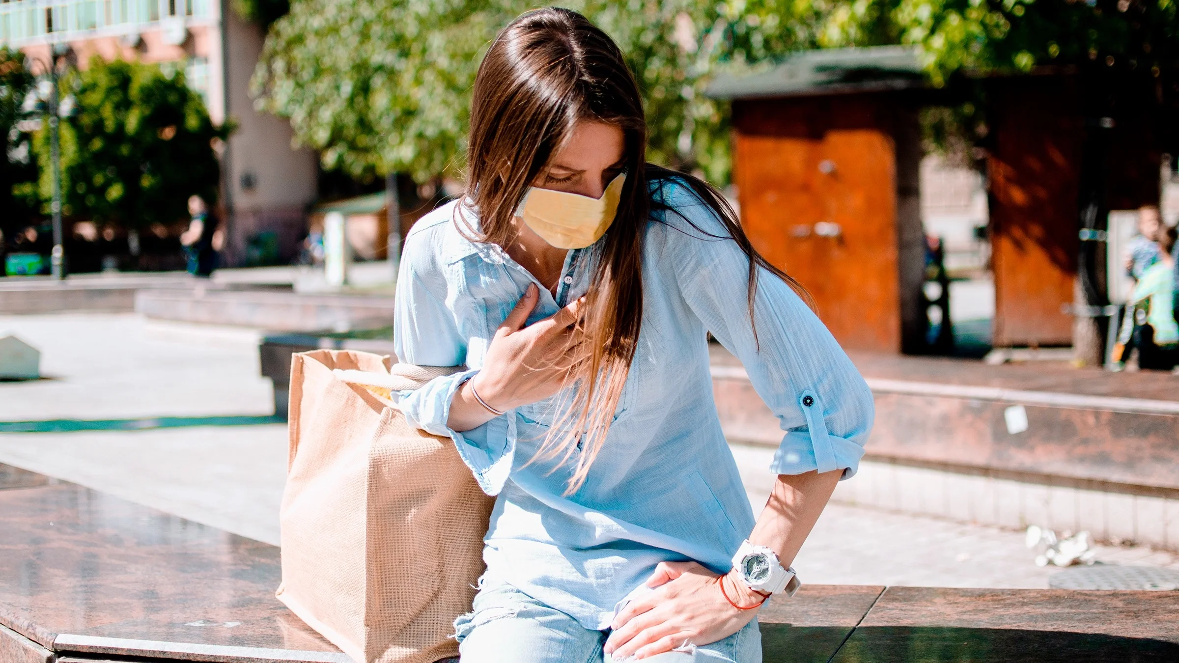 Young female wearing a yellow face mask having trouble breathing and holding her chest while resting in city street