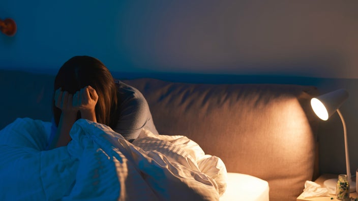 A person with a headache in bed, with pills on their nightstand. 