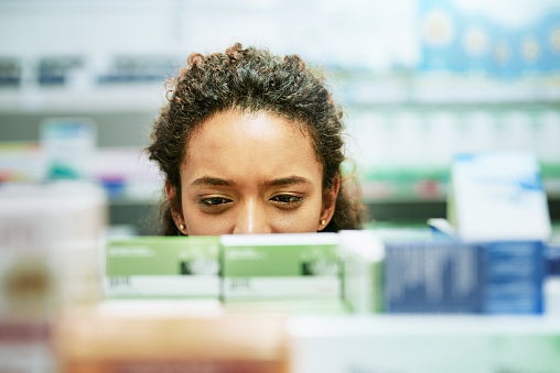 Close-up of young woman looking medications on the shelf at the pharmacy, shot looking at her over the aisle with only her nose and eyes in view.