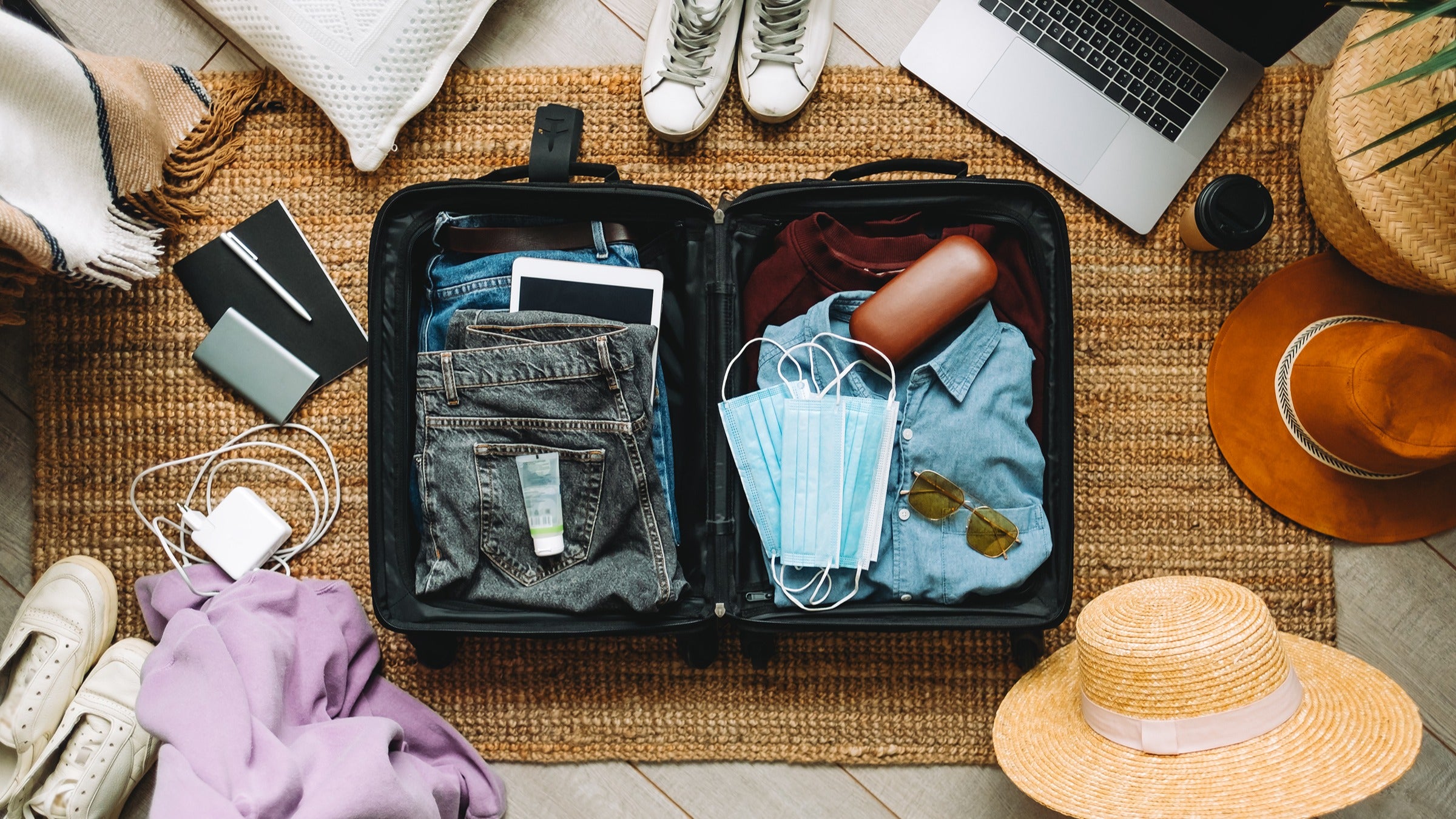 Overhead shot of packing a suitcase. The suitcase is full of clothes, face masks, and sunglasses. There are hats, shoes, and other miscellaneous things to pack surrounding the suitcase on the floor.