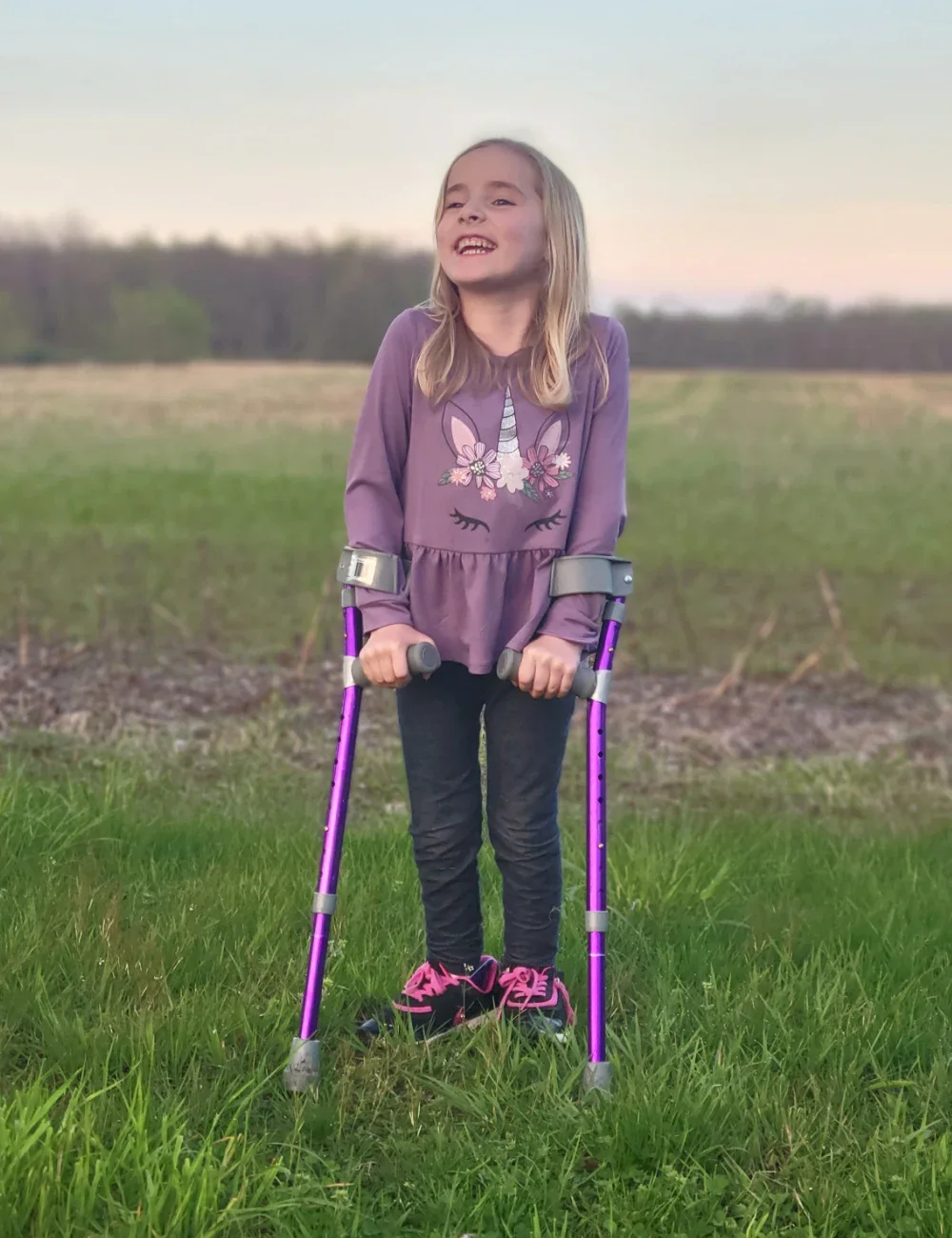 Sophia Kleeh, age 7, is pictured in a field wearing a purple shirt and walking with her purple forearm crutches.