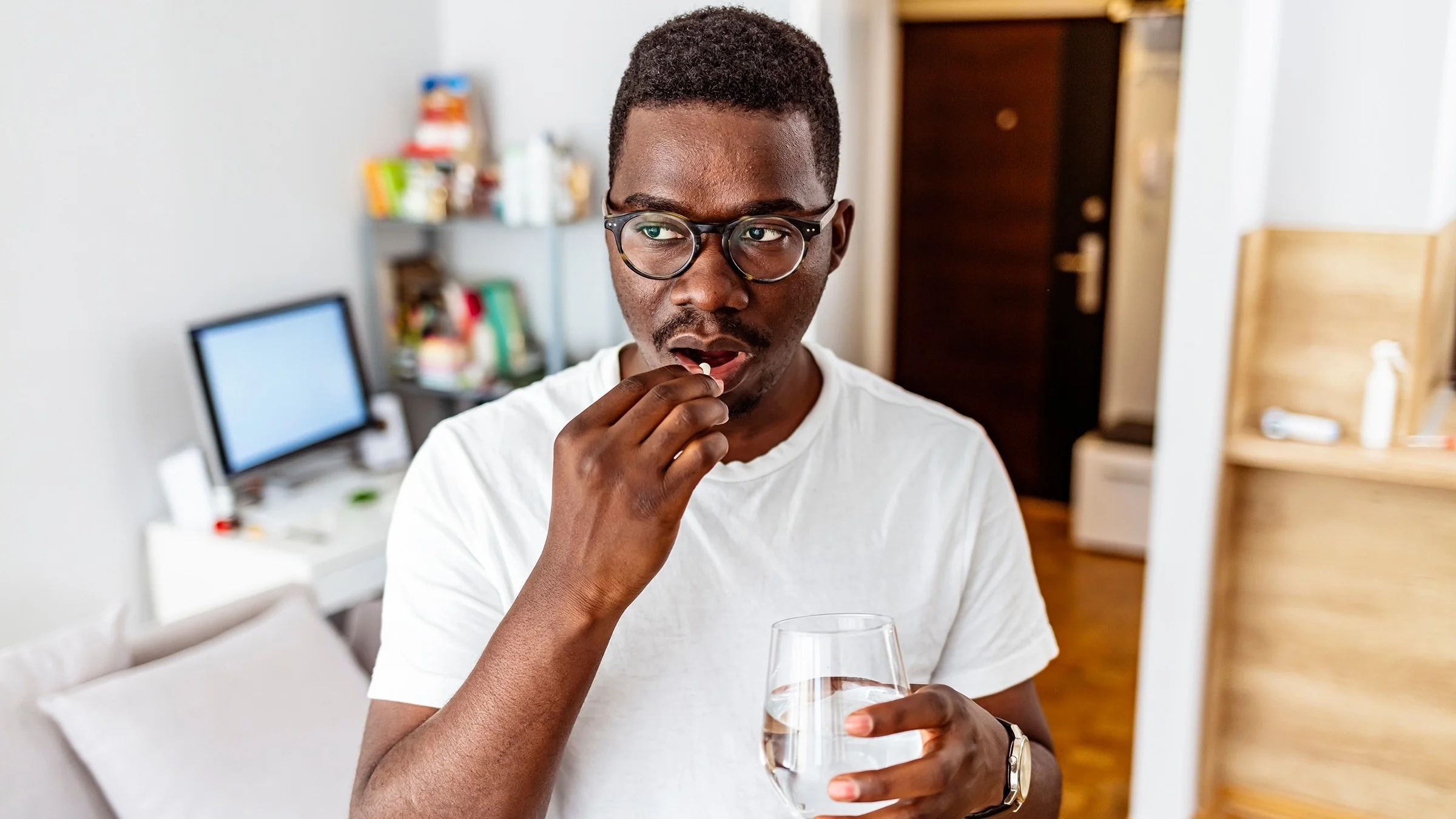 Portrait of a man taking a small white pill with a glass of water in his apartment.