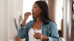 Portrait of a young pregnant woman taking a white oblong pill. She is wearing a white shirt with a denim button up over it.
PeopleImages/iStock via Getty Images
