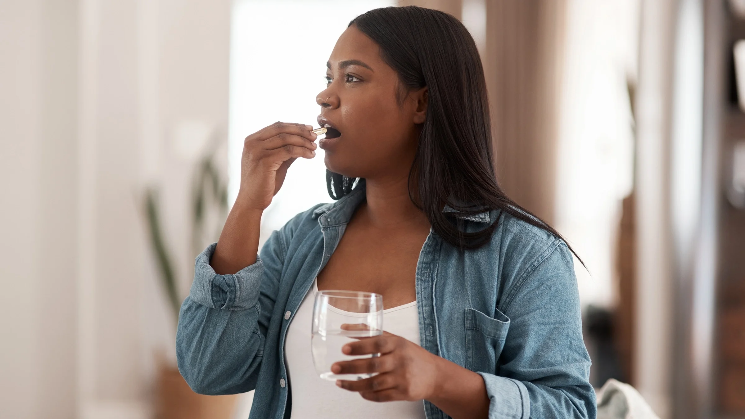 Portrait of a young pregnant woman taking a white oblong pill. She is wearing a white shirt with a denim button up over it.