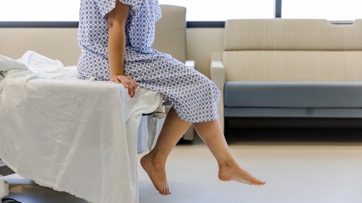 A patient in a hospital gown sits on the end of a hospital bed.