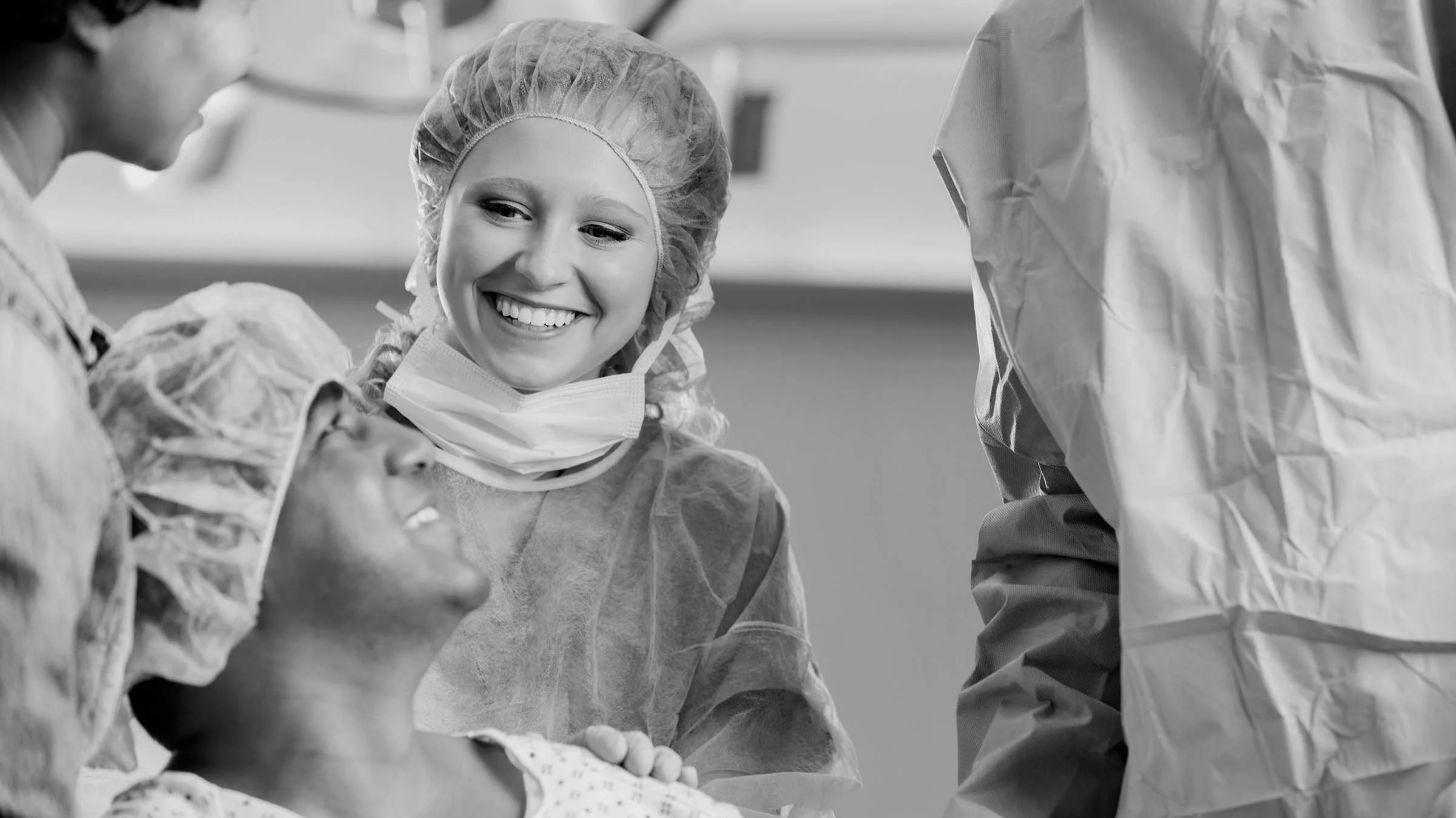 Black-and-white image of nurses helping a patient into a surgery room. The patient is looking up at one of the nurses and smiling from a wheelchair.