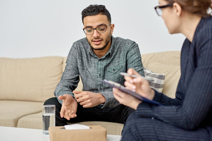 Young man talking to a therapist in a simple office, he's sitting on a tan couch with a glass of water in front of him.