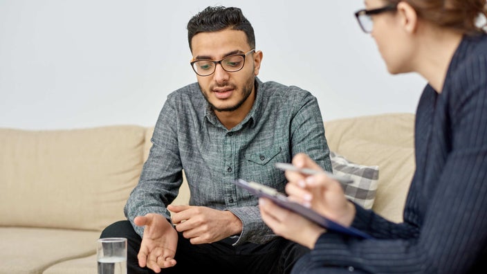 Young man talking to a therapist in a simple office, he's sitting on a tan couch with a glass of water in front of him.