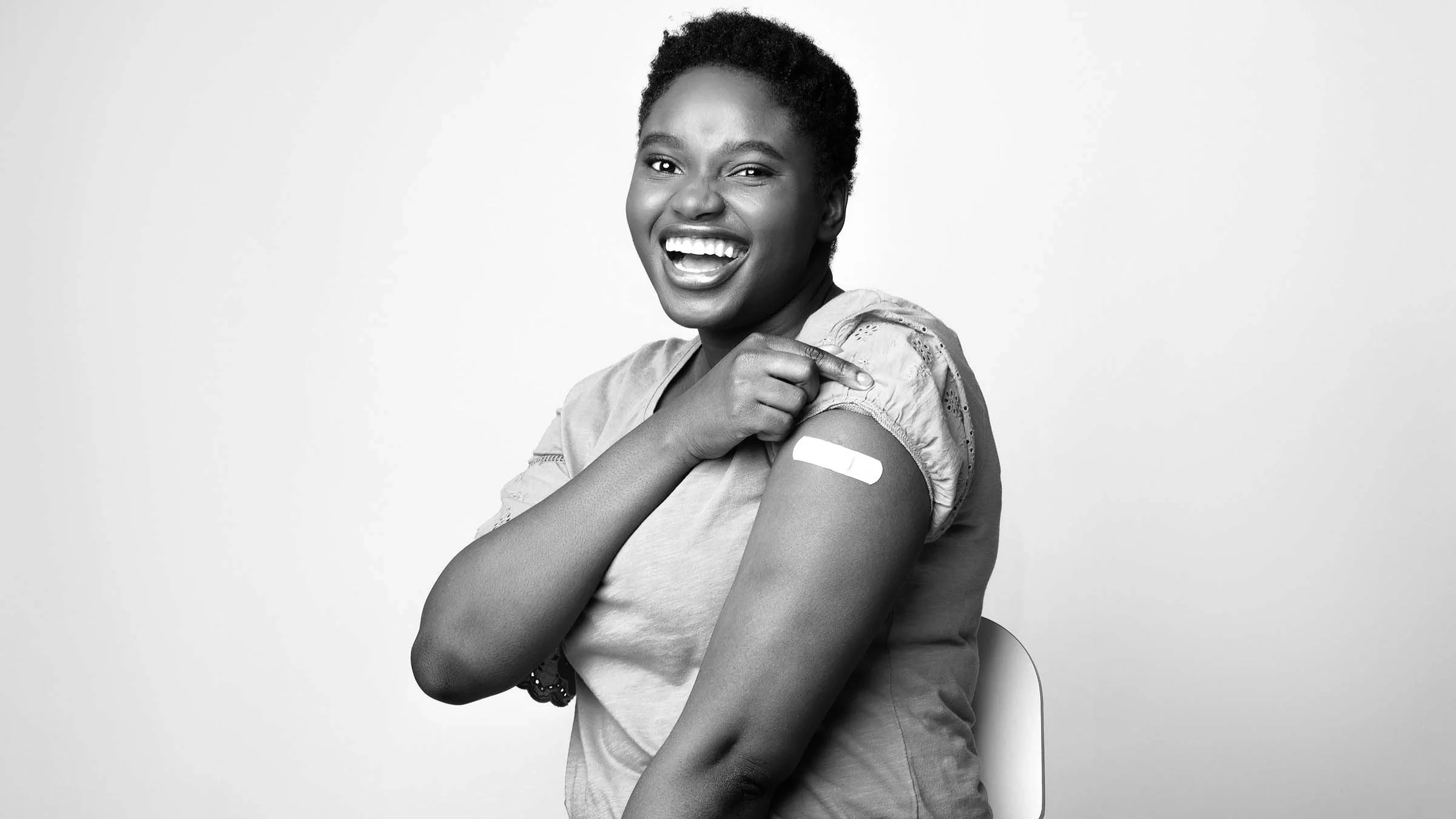 Black and white portrait of a smiling woman showing off her band-aid after the COVID-19 vaccine. She is sitting down in a chair in a studio setting.
