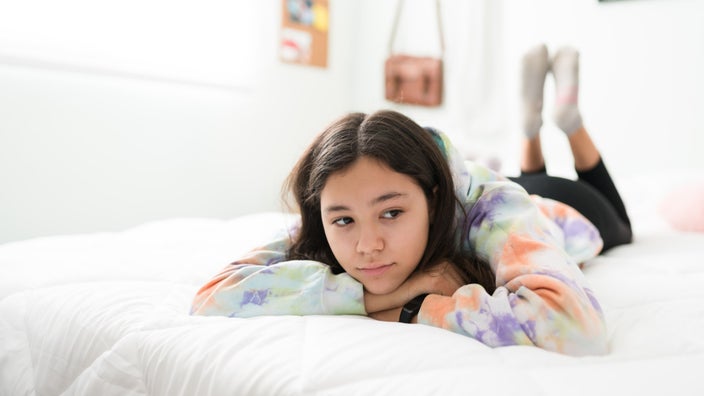 Sad looking teenage girl laying on her bed in a tie dye sweatshirt. She's laying on her stomach with her chin resting on her hands.