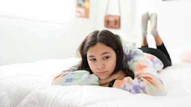Sad looking teenage girl laying on her bed in a tie dye sweatshirt. She's laying on her stomach with her chin resting on her hands.
antoniodiaz via Shutterstock