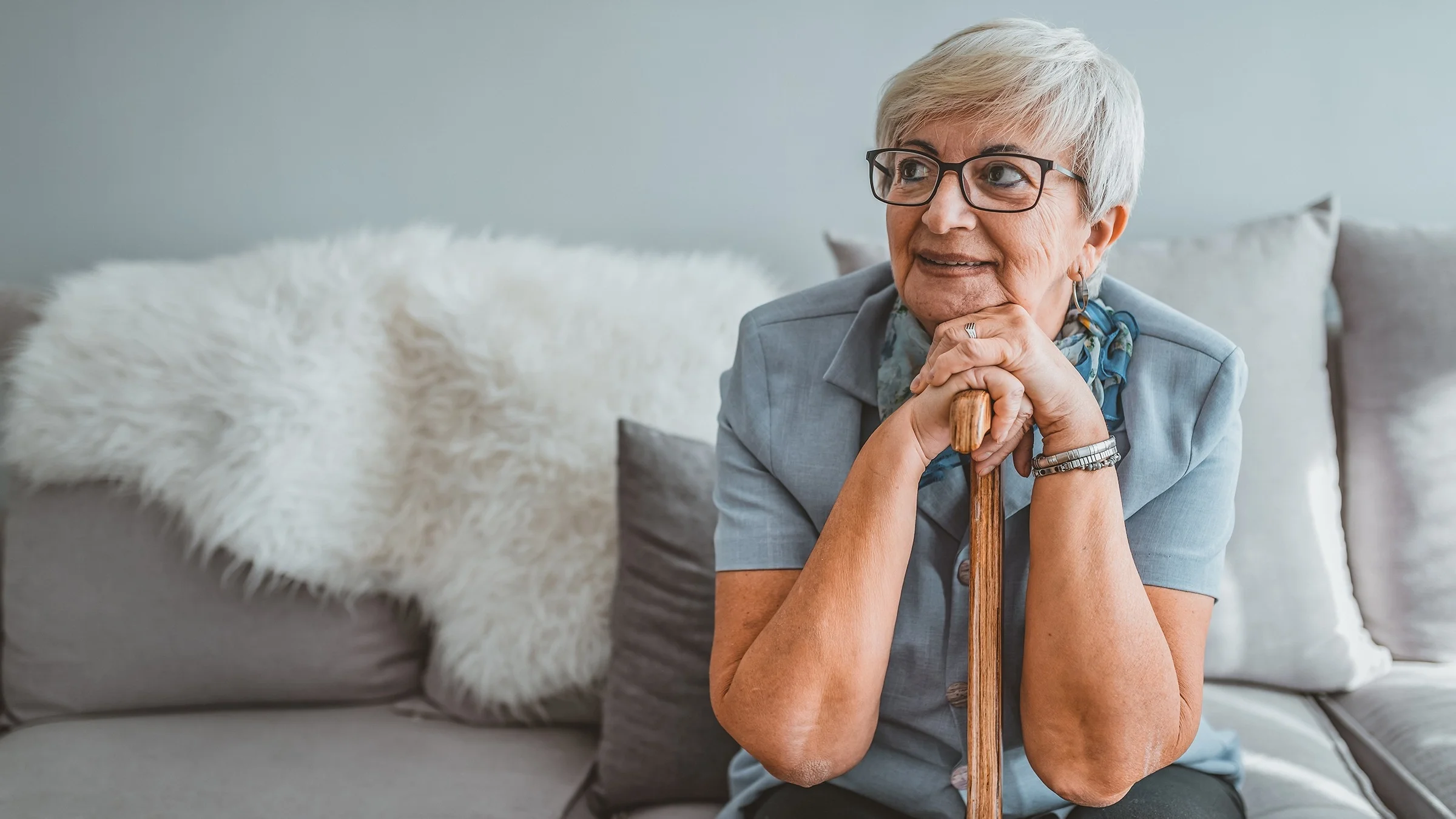 Portrait of an elderly woman sitting on the couch and leaning forward resting her hands and chin on her wooden cane.